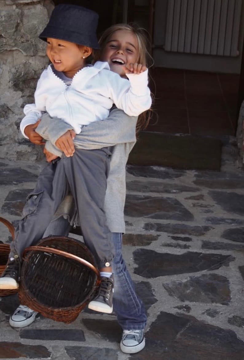 Two children laughing outside a stone house, one carrying the other, with a wicker basket